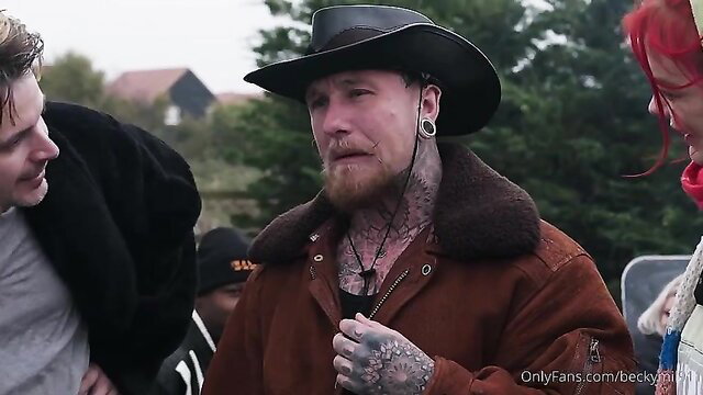 Tattooed bearded man in cowboy hat and fur coat with redhead at outdoor event