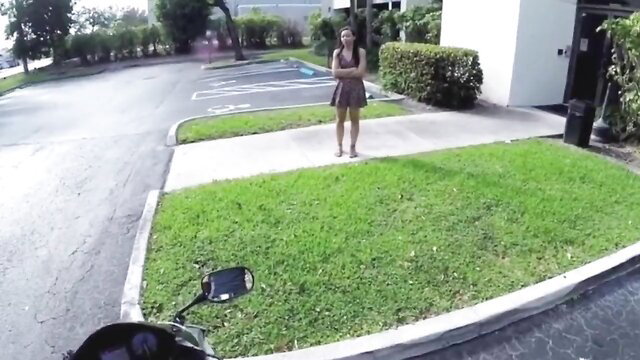 Black teen in short dress stands by motorcycle in outdoor parking lot POV