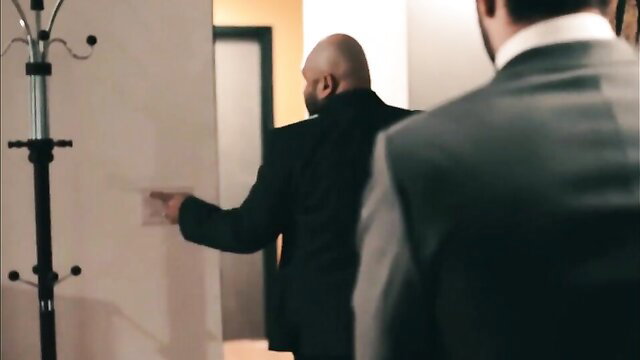 Bald muscular man in suit points seductively with suited man behind in hallway
