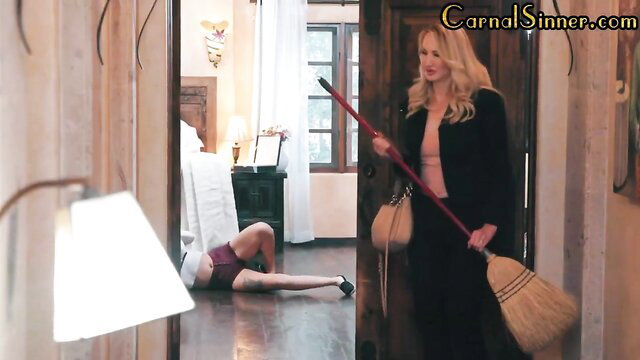 Blonde seductress in black coat holds broom over curvy BFF on floor