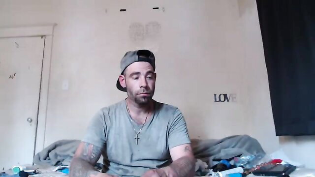 Tattooed white guy in cap sits on bed in amateur solo strip tease