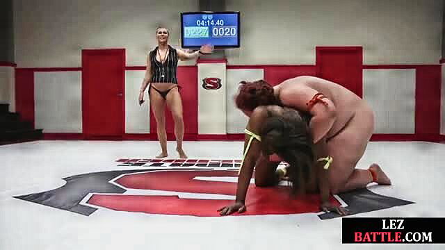 Blonde ref stands over black BBW wrestler pinning nude opponent on mat