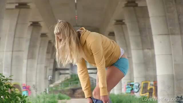 Blonde Violeta in yellow hoodie and blue shorts bends over under bridge