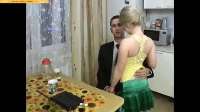 Blonde woman in yellow top and green skirt straddles suited man in kitchen
