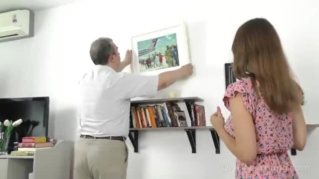 Kecy Hill in pink dress watches man hang picture on shelf