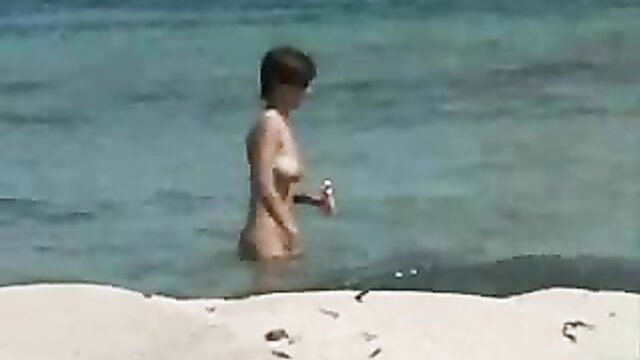 Nude Asian woman with short black hair holding bottle in shallow sea at nudist beach