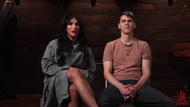 BDSM shemale in robe dominates young man in wood dungeon