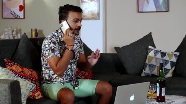Indian man in colorful shirt shorts on phone with whiskey on couch