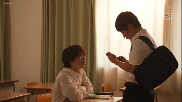 Two Japanese schoolboys in classroom, one showing phone to seated friend