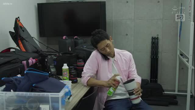Asian man on phone in studio, casual pink shirt, surrounded by camera gear