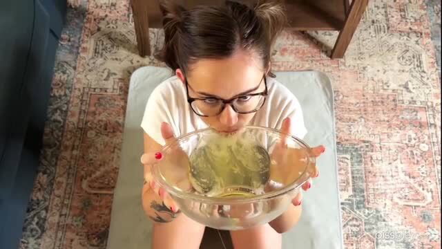 Tattooed brunette in glasses holds bowl of piss close to mouth in fetish scene