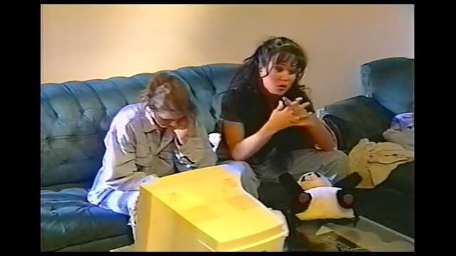 Two dark-haired women on blue couch reacting to retro video game, clothed softcore scene