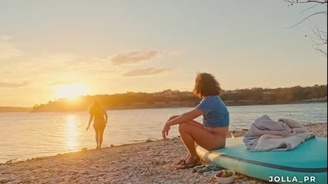 Curly-haired woman in blue crop top by water at sunset with paddleboard