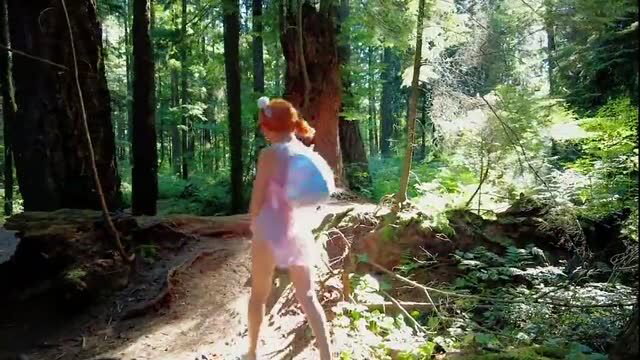 Redhead in sheer pink dress walks forest trail with fairy wings
