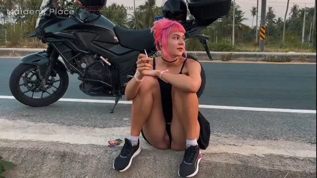 Pink haired woman squatting roadside beside black motorcycle in bodysuit