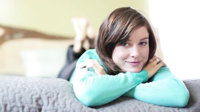 Zoey smiles playfully on bed in teal sweater, showing her technique