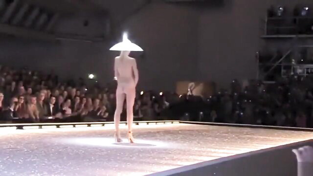 Nude model with conical hat struts seductively on glitter runway
