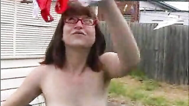 Nude woman with glasses holds panties over head in backyard tease