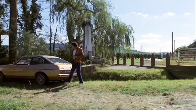Vintage 70s guy in jeans walks by yellow Ford Escort near bridge in field