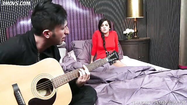 Caroline Ray in red dress on bed as guy plays guitar seductively
