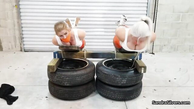 Two busty blondes in orange bikinis bound on tire racks BDSM pose