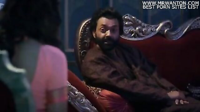 Indian man in robe feeds woman laddoo on ornate sofa