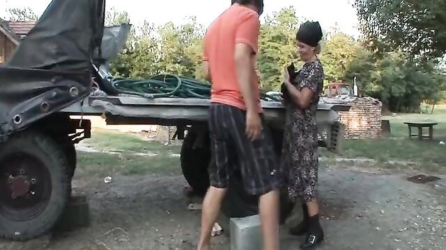 Old farmer wife in floral dress talks to bare-chested man by truck