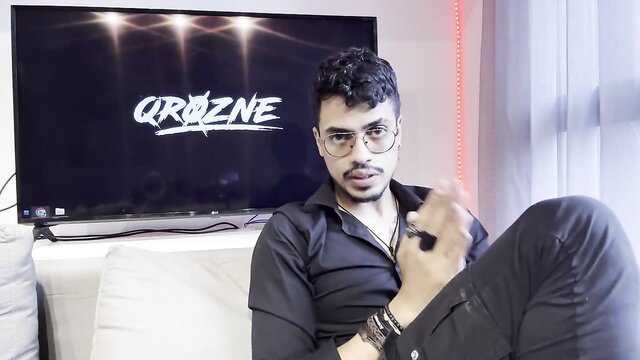 Curly-haired Arab man in black shirt poses on couch for QROZENE adult video thumbnail