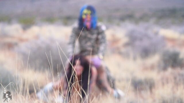 Drea Alexa with blue hair sits in desert, camo top skirt legs spread