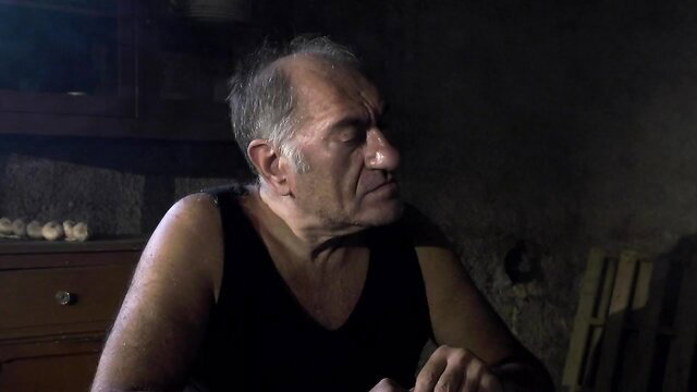 Old man Eddie in tank top, intense gaze in dim room