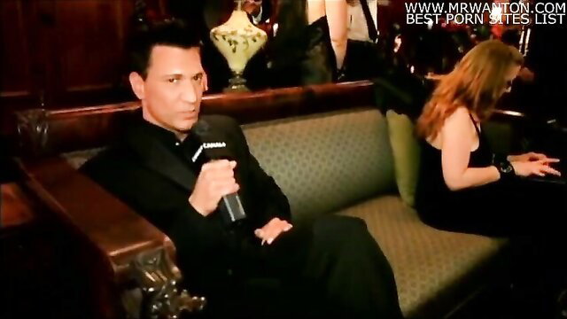 Man in tuxedo interviews redhead woman in elegant dress on green couch